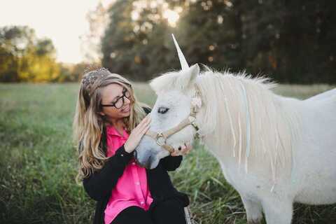 Cassidy sitting in a hot pink shirt and black cardigan, snuzzling a miniature pony turned unicorn
